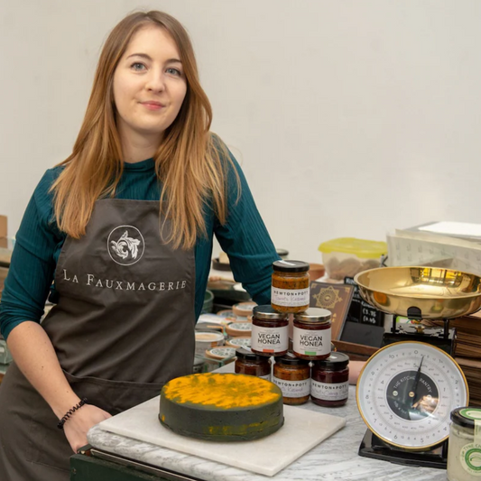 A woman wearing a dark green top and a brown apron with the logo “La Fauxmagerie” stands behind a counter displaying vegan cheese, jars labeled “Vegan Honea” and “Newton & Pott,” and a vintage-style weighing scale.
