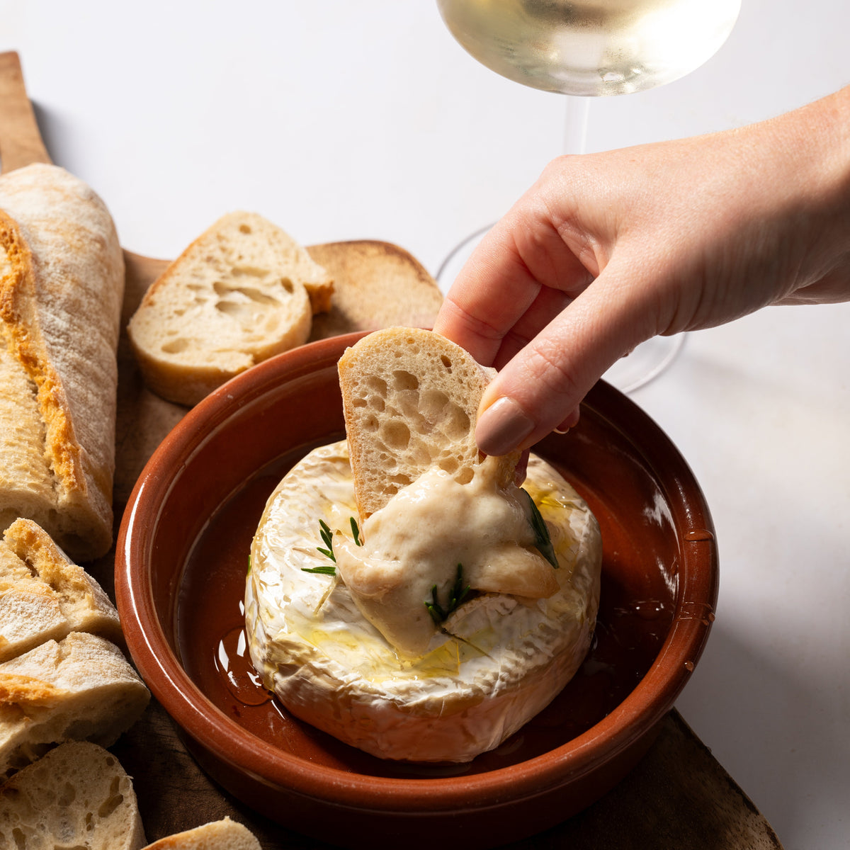 A hand dipping a piece of rustic bread into a terracotta bowl filled with creamy baked camembert, olive oil, and herbs, with sliced bread and a glass of white wine on a wooden board.