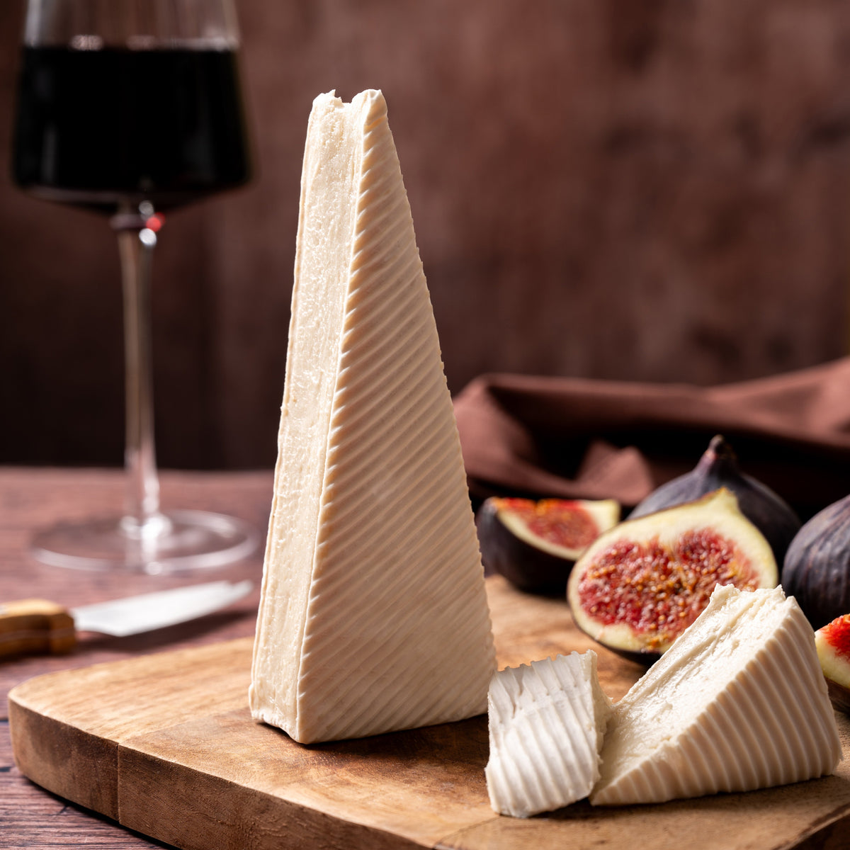 A wedge of vegan brie on a wooden cutting board, with sliced figs and a glass of red wine in a rustic brown setting.