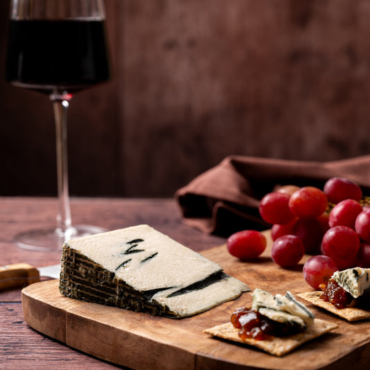 A styled food scene on a wooden board with a wedge of blue cheese, crackers topped with jam and cheese, red grapes, and a glass of red wine against a dark brown rustic background.