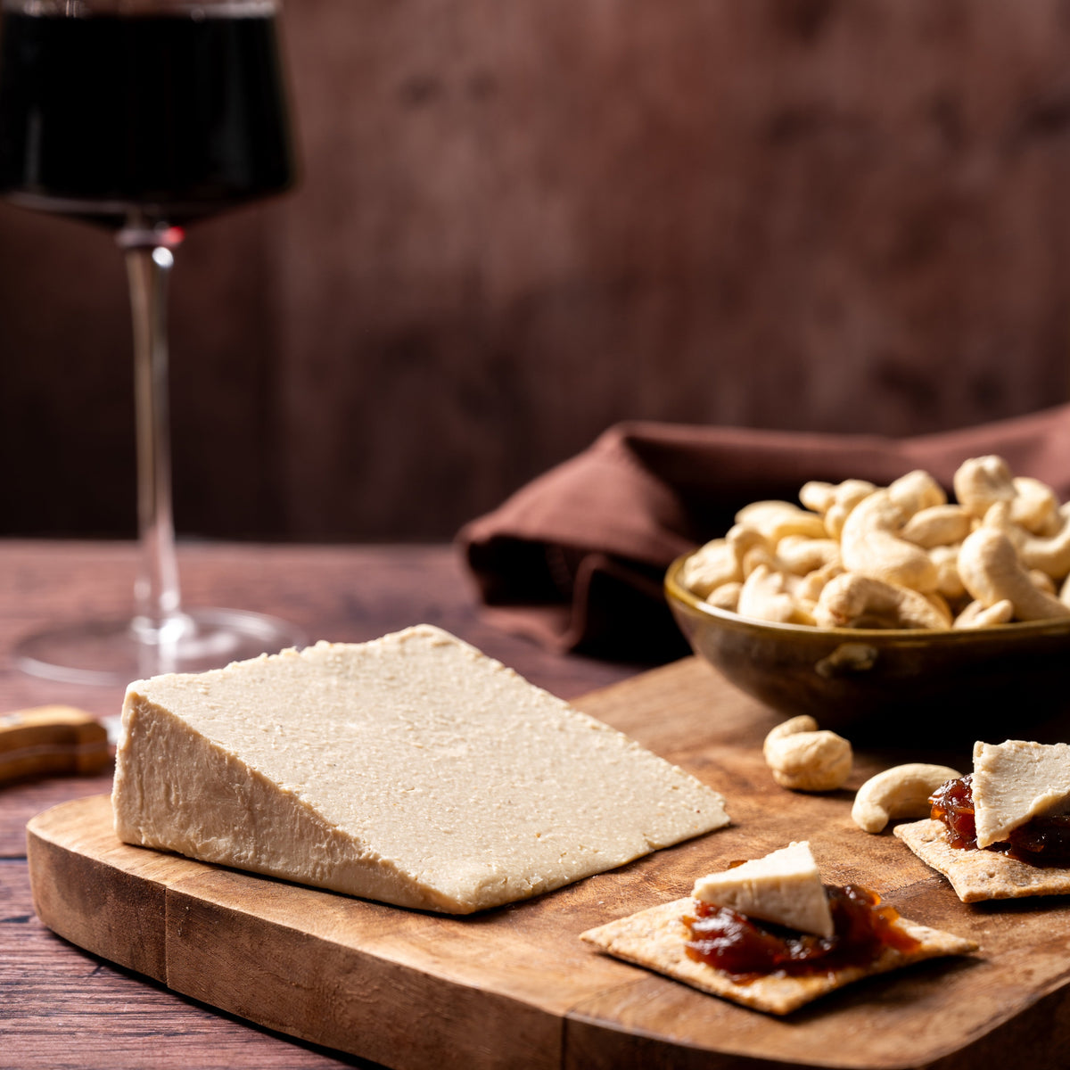 A styled food scene on a wooden board with a large rectangular block of pale cheese, crackers topped with cheese and jam, a bowl of cashews, and a glass of red wine in the background on a dark wooden table.
