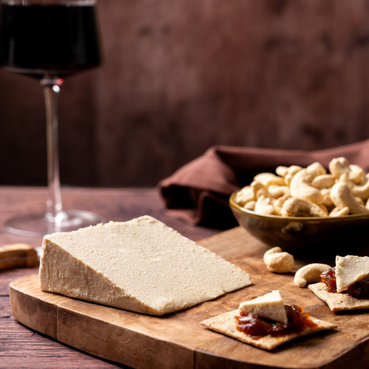 A styled food scene on a wooden board with a large rectangular block of pale cheese, crackers topped with cheese and jam, a bowl of cashews, and a glass of red wine in the background on a dark wooden table.