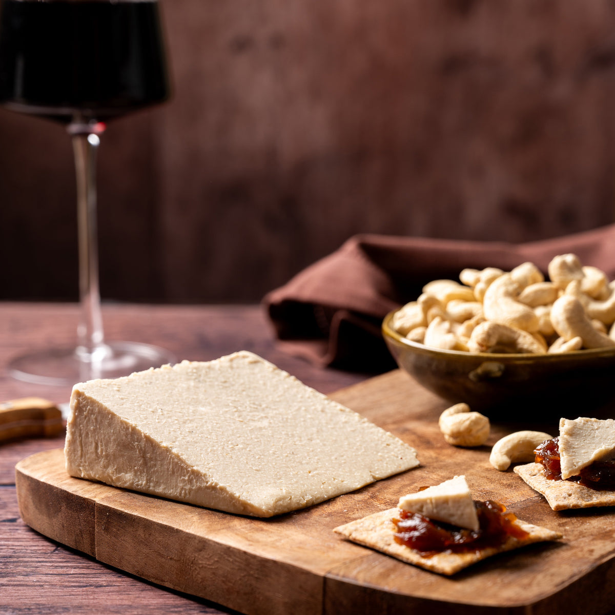 A styled food scene on a wooden board with a large rectangular block of cheese, crackers topped with cheese and jam, a bowl of cashews, and a glass of red wine in the background on a dark wooden table.