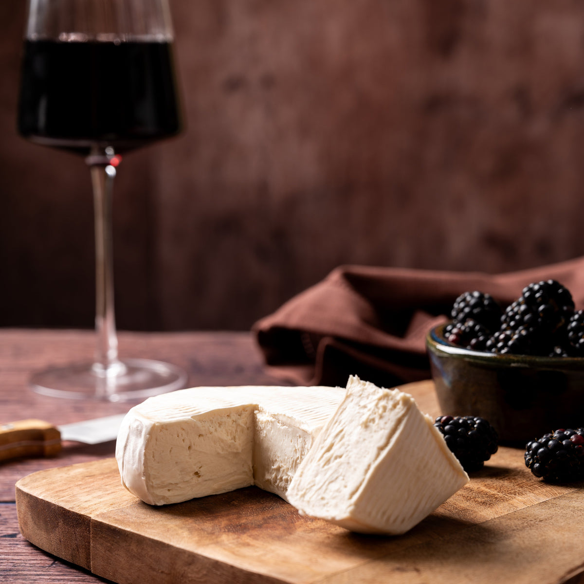 A wedge of soft white cheese camembert style on a wooden cutting board, with a glass of red wine and a bowl of blackberries in a rustic setting.