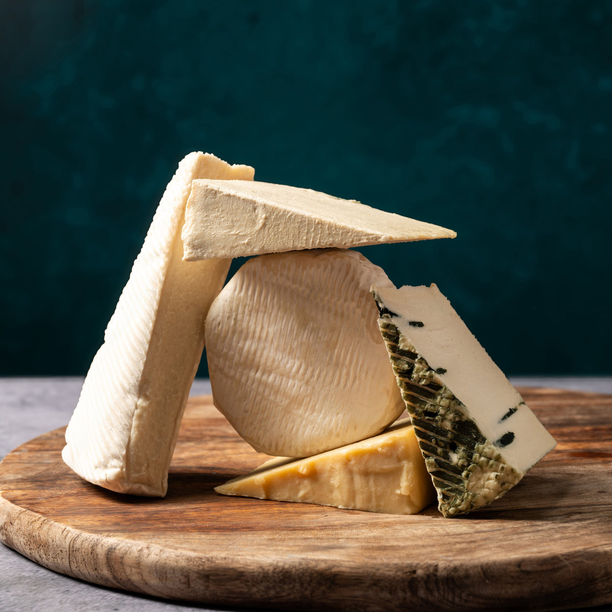 An assortment of artisanal cheeses, including a round wheel of soft cheese, wedges of hard cheese, and a blue cheese slice with green mold, arranged on a round wooden cutting board against a dark teal background.