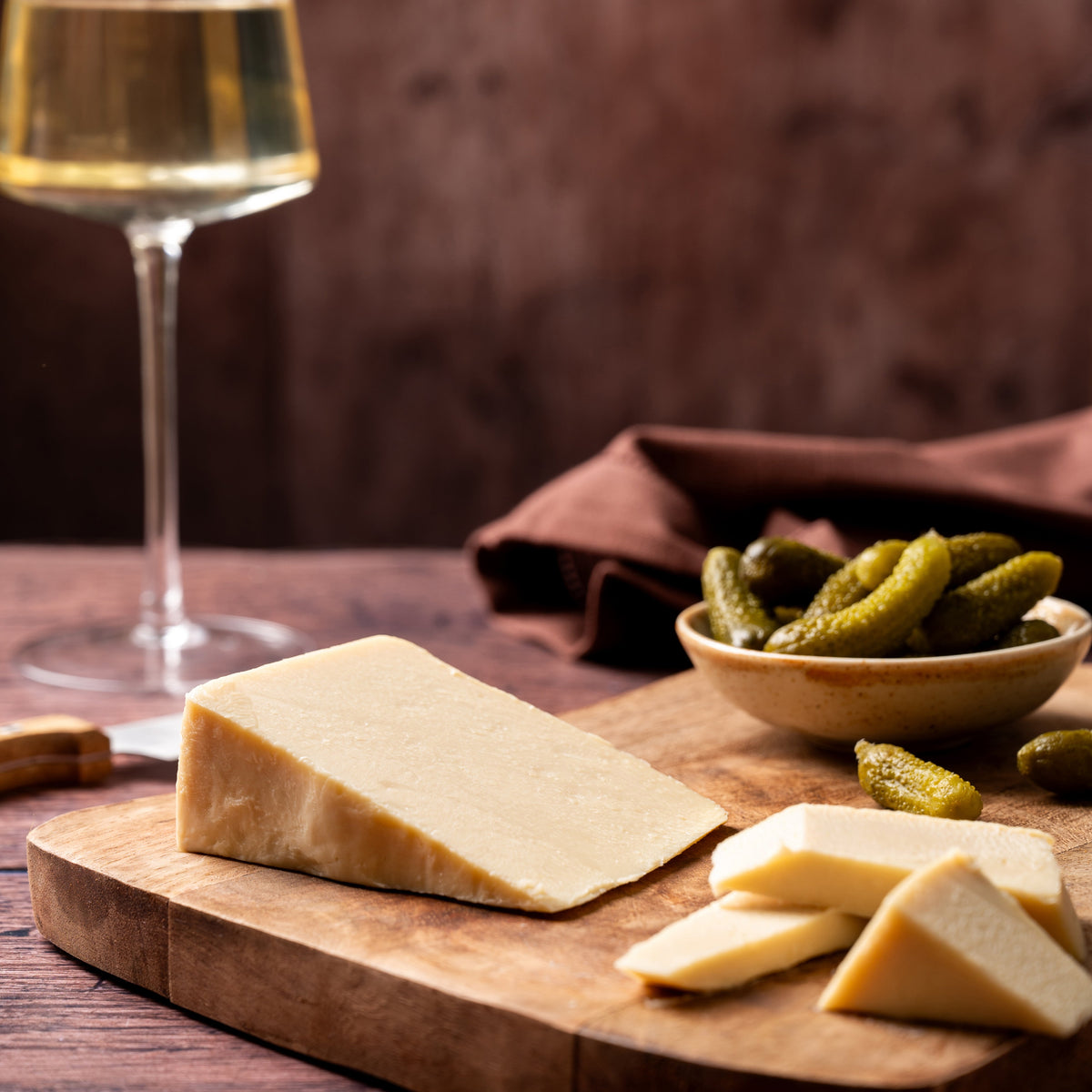A styled food scene on a wooden board with a wedge and slices of pale cheese, a small bowl of pickles, and a wine glass in the background against a dark rustic backdrop.