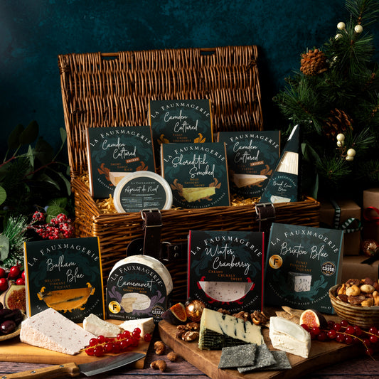 A curated cheese gift basket arrangement on a rustic wooden table, featuring a wicker basket filled with packaged cheeses, sliced cheeses on a board, nuts, and small decorative holiday elements against a dark teal background.