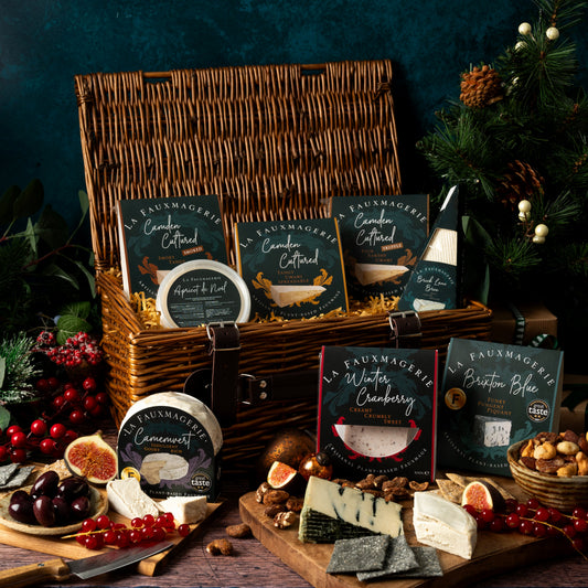 A wicker picnic basket filled with packaged gourmet foods and cheese, arranged on a rustic wooden table with sliced cured meats, crackers, figs, nuts, and red berries against a dark teal background with evergreen branches.