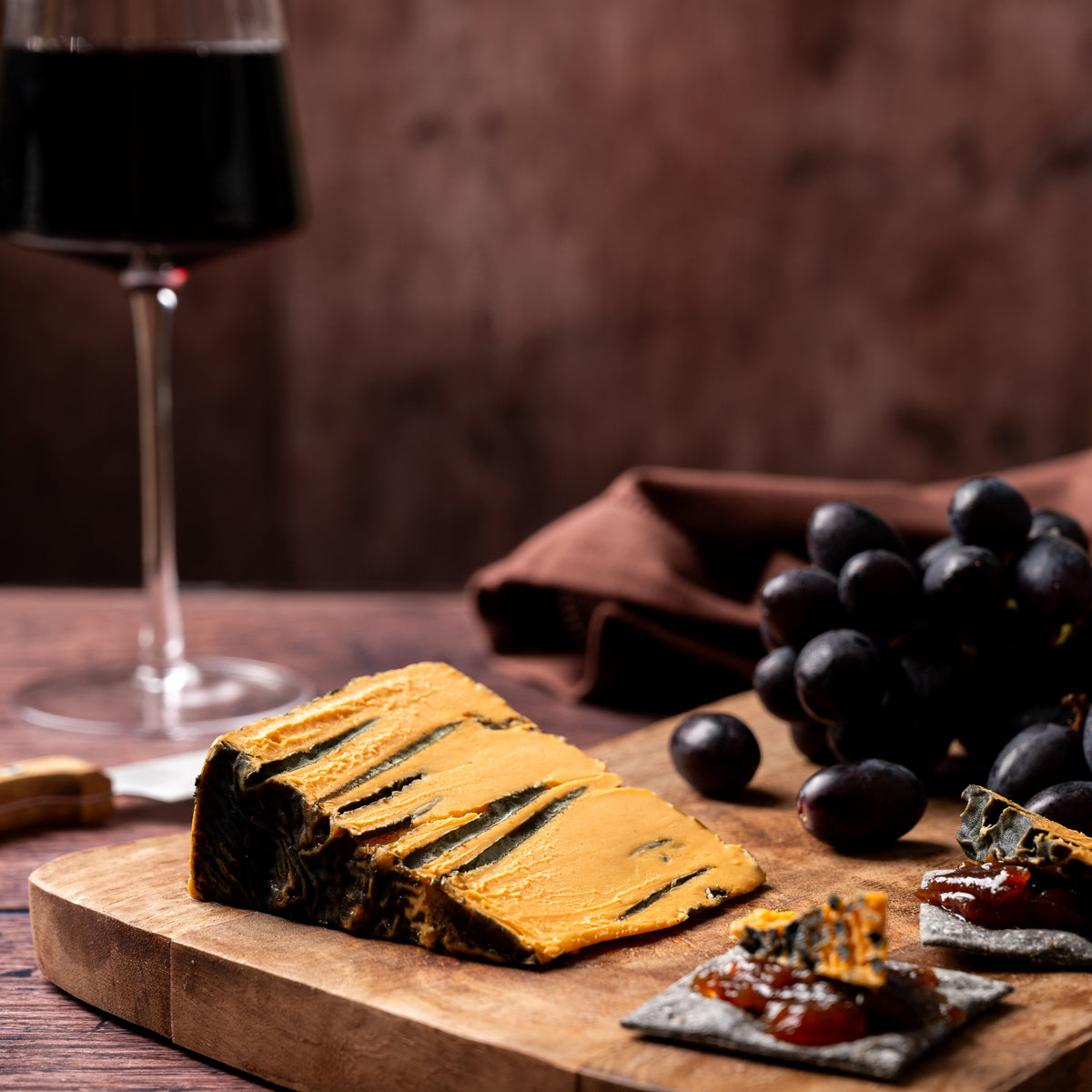 A styled food scene on a wooden board with a wedge of balham blue orange vegan cheese, dark grapes, small appetizer crackers, and a glass of red wine against a dark brown rustic background.